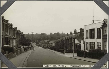 Black and white photograph of an urban street scene featuring rows of terrace houses on either side of a tarmac road that gently slopes and curves into the distance. To the left, two-storey houses with canted ground floor bays and small front gardens front onto the pavement. Opposite, a distinct, two-storey modernist building topped with a clock and flag pole stands on the corner of the road and an access road. Further along the right side of the road are terraced houses with gables facing the street. Two cars are parked on the road in the far distance and a woman in a long coat and carrying a bag crosses the access road. A label applied to the bottom-right of the photograph reads: 'East Barnet. Brunswick Park Road, part of which is obscured by one of four corner photograph album mounts. 