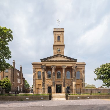 Front view of a sandstone church with darkened features, columns, pediment and square tower. 