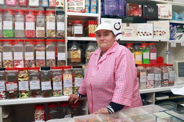 A photograph of a woman wearing a pink and white checked shirt and a white cap, behind the counter in a sweet shop. Jars of sweets line the wall behind her.