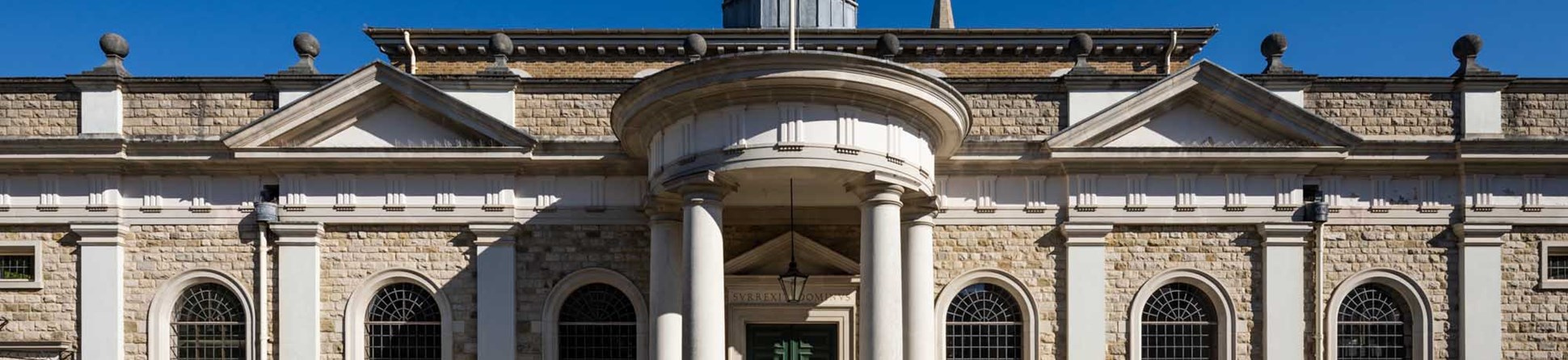 The exterior of Brentwood Cathedral shows a cobbled and paved circular external flooring. A rounded porch with pillars and a long line exterior wall with semi-circular leaded windows and two arches, with a decorative hexagonal tower.