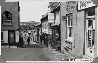 Black and white photograph of an urban street scene. The view looks along a curving street that drops downhill. The right side of the street has two-storey buildings that front onto a narrow cobbled path. To the left foreground, the road widens in front of a two-storey building with a first-floor arched sash window. Many of the buildings on the right side are shops carrying various signs and advertisements. A lady wearing a hat and carrying a shopping bag steps from one of them. The buildings are variously clad in render and tiles. A label applied to the bottom-right of the photograph reads: 'Launceston. Northgate Street', part of which is obscured by one of four corner photograph album mounts.