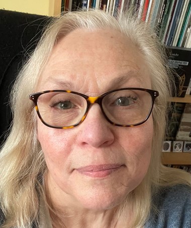 Photograph showing close up of woman’s face with light hair and black frames glasses. 