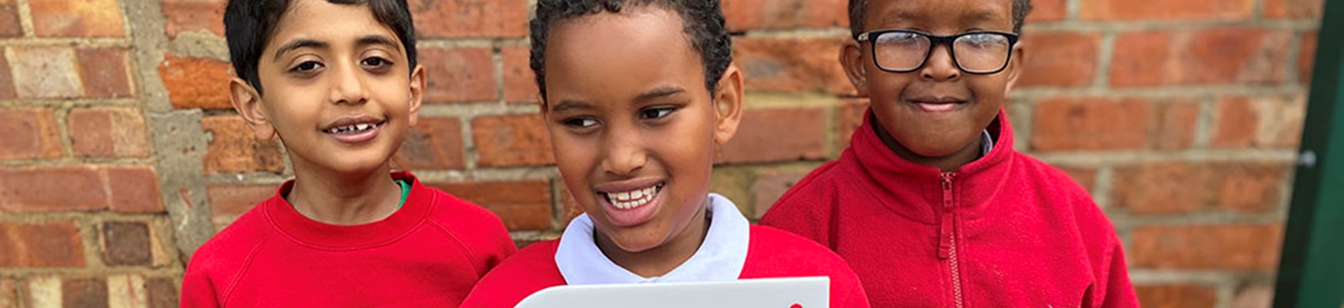 Three children in red school jumpers stand in front of a brick wall, holding up a plaque that reads "Historic England: Heritage Schools Award".