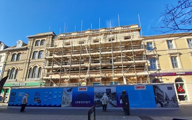 Stone-built period building shrouded in scaffolding with information boarding at ground level. Pedestrians passing by are reading the information panels.